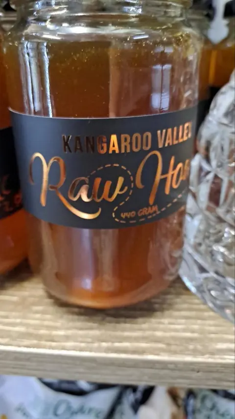 Close-up of a 440 g jar of Kangaroo Valley Raw Honey (black and copper label) on a timber shelf beside cut glass; soft focus