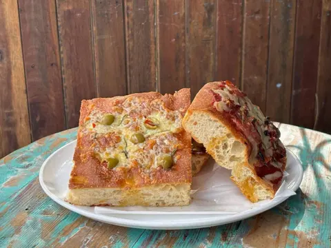 Squares of house sourdough pizza bread (olive and cheese; salami and tomato) on a white plate on a worn turquoise table against a dark timber wall