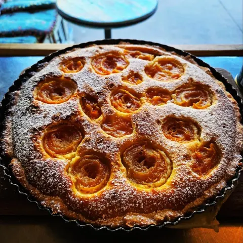 Warm peach frangipane pie dusted with icing sugar in a fluted tin, turquoise verandah table behind