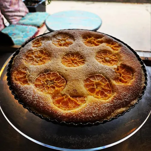 Mandarin frangipane tart (whole mandarin halves set in almond sponge) on a glass cake stand, turquoise verandah table behind