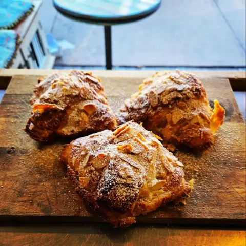 Three almond croissants dusted with icing sugar on a worn timber board, turquoise verandah table behind
