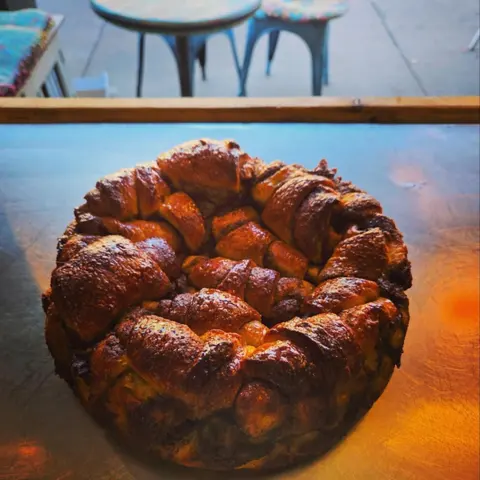 Round cinnamon babka, glossy pull-apart crown on the steel bench, blue metal chairs on the verandah behind