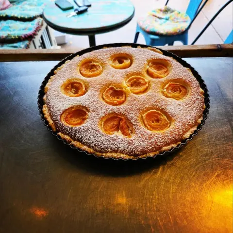 Apricot frangipane tart dusted with icing sugar on the steel bench, turquoise verandah table and floral chair cushion behind
