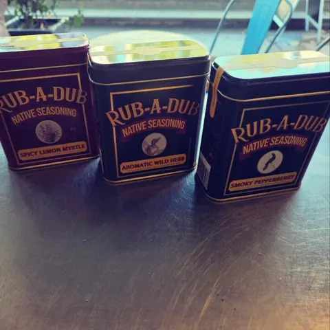 Three tins of Rub-A-Dub Native Seasoning (Spicy Lemon Myrtle, Aromatic Wild Herb, Smoky Pepperberry) on the steel bench, verandah behind