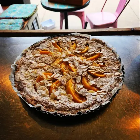 Gluten-free peach and almond tart in a fluted tin on a steel bench, pink metal cafe chairs on the verandah behind