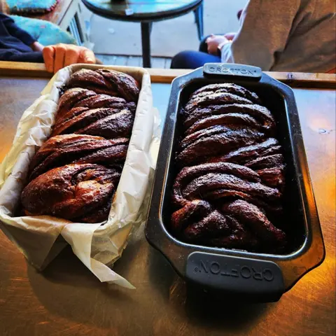 Two loaves of chocolate rollada (babka) in tins on a cafe table, customers' hands in background
