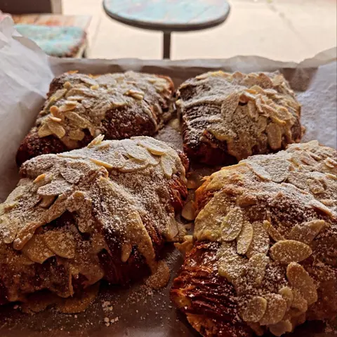 Four house-made almond croissants, flaked almonds and icing sugar, on baking paper, cafe table behind