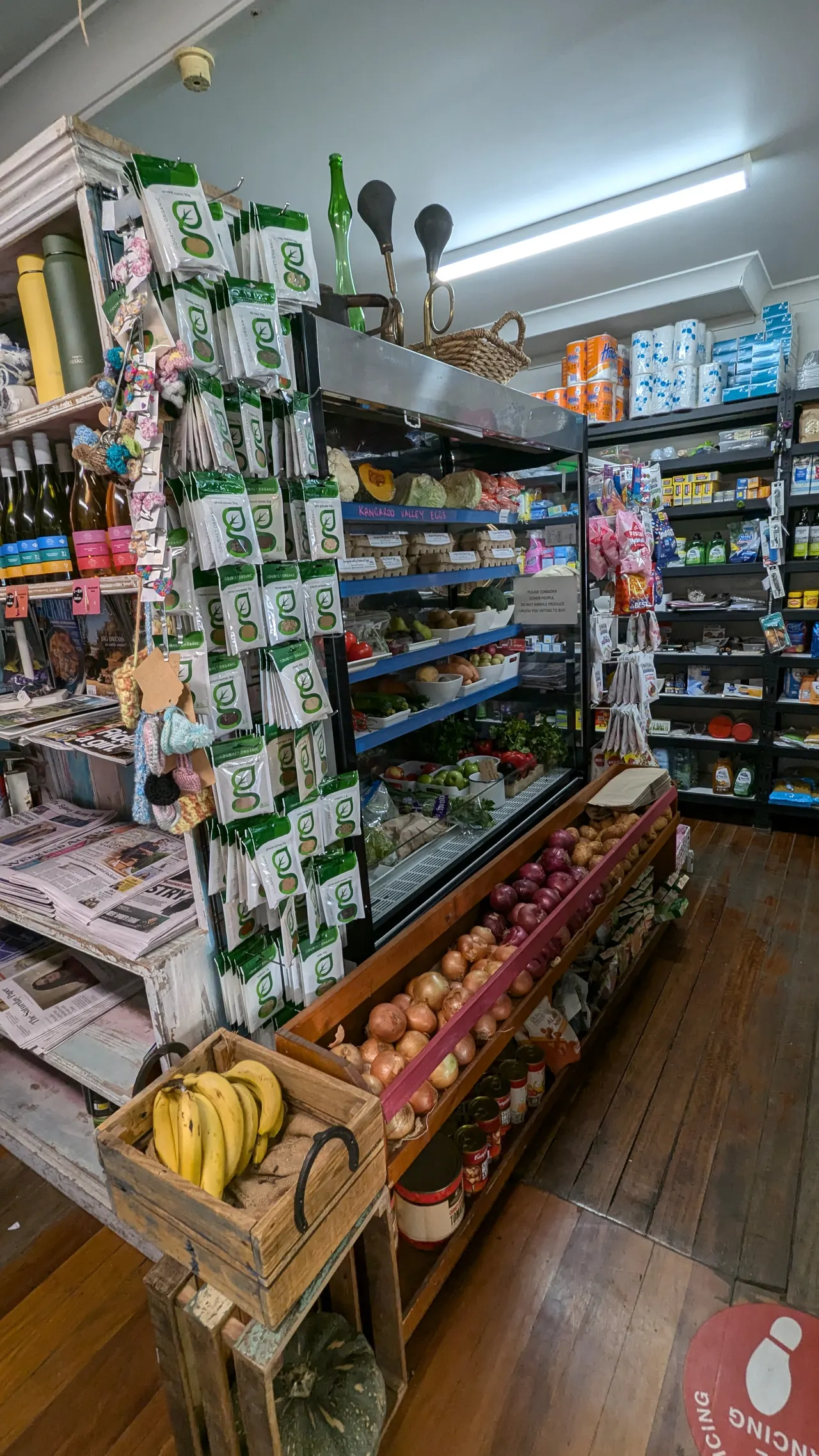 An aisle of the store: newspapers and magazines on the rack, greeting cards, bananas and crates of produce.
